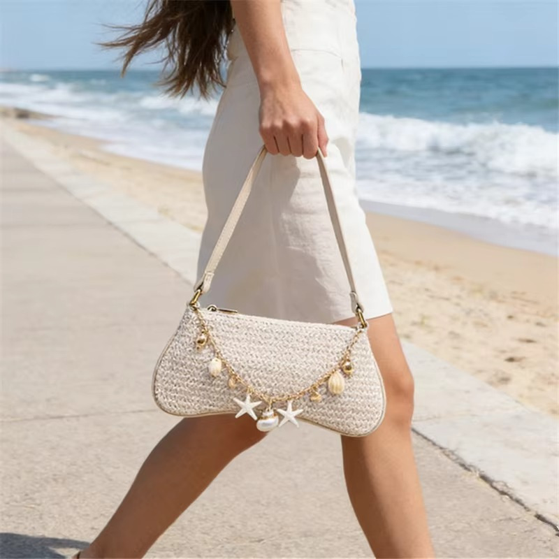 Sac en paille baguette beige avec bandoulière et chaînette ornée de breloques coquillages porté sur la plage