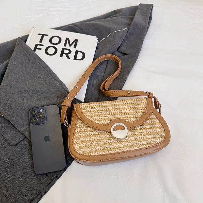 Sac en paille bandoulière avec rabat arrondi et bordure cuir marron clair posé sur un vêtement gris à côté d'un smartphone noir et d'un livre Tom Ford