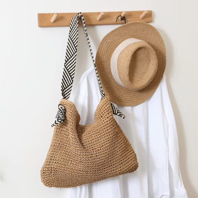 Sac en paille crochet beige avec bandoulière rayée noire et blanche modèle Bertille suspendu à un porte-manteau en bois à côté d'un chapeau en paille et d'une veste blanche
