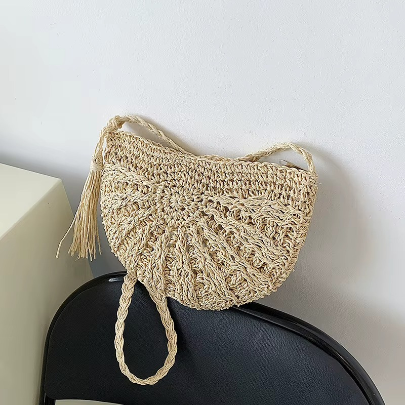 Sac en paille demi-cercle couleur amande avec pompon et bandoulière tressée posé sur un dossier de chaise noire