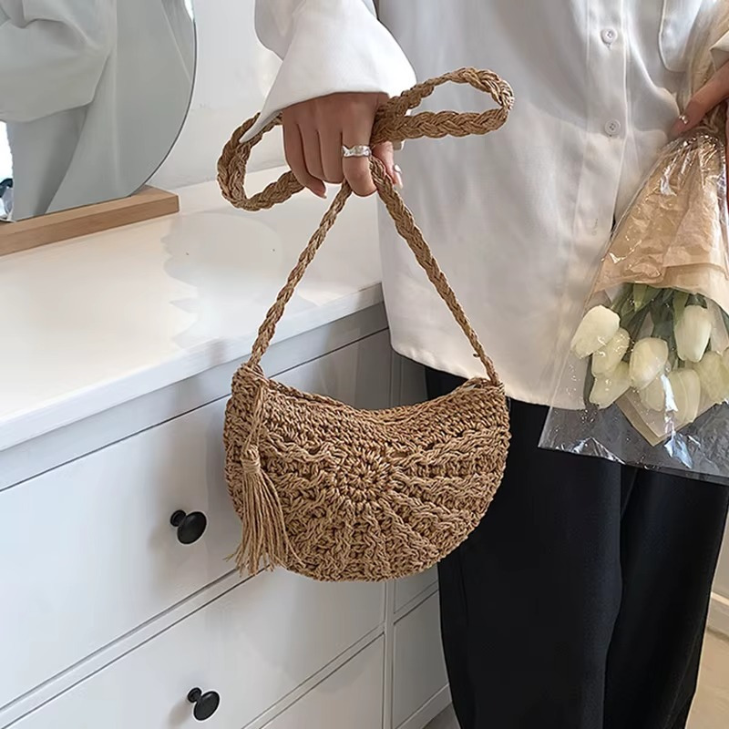 Sac en paille demi-cercle couleur naturelle avec pompon et bandoulière tressée tenu à la main