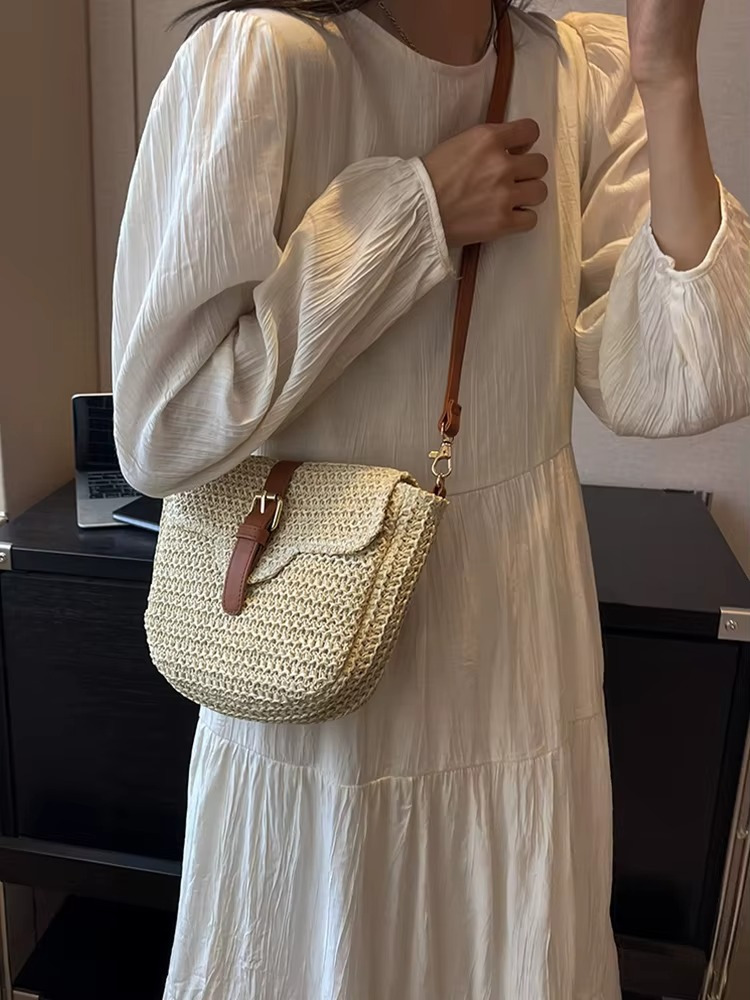 Sac en paille couleur naturelle avec rabat et boucle en cuir marron porté en bandoulière par une femme en robe blanche