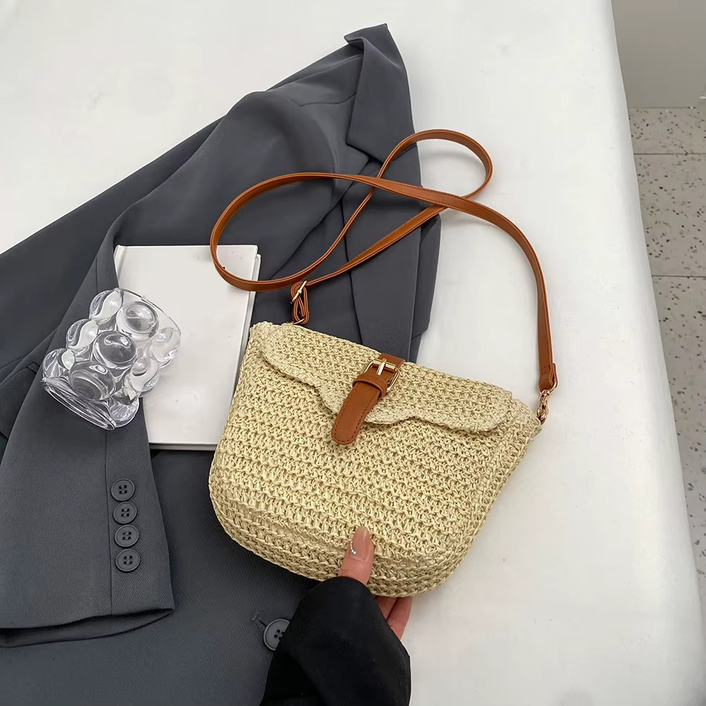 Sac en paille couleur naturelle avec rabat et boucle en cuir marron modèle Ophélie, porté à la main sur un fond gris avec veste et verre