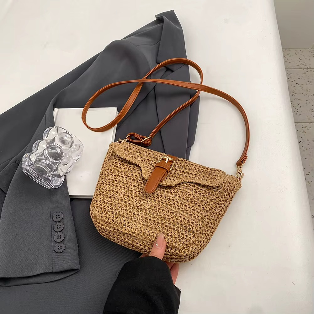 Sac en paille avec rabat à boucle en cuir marron, porté main, sur fond de veste grise