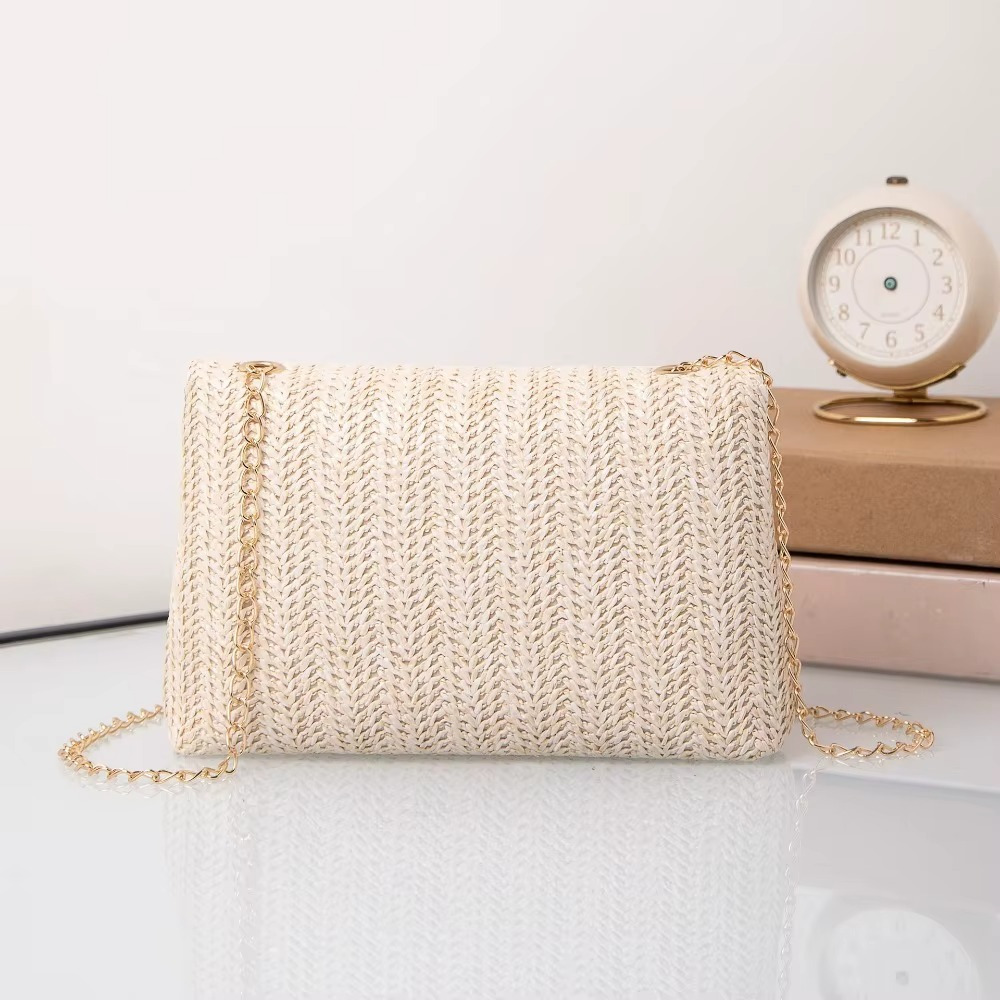 Sac en paille beige avec rabat et fermoir chaîne modèle Angélique posé sur une surface blanche avec une horloge en arrière-plan