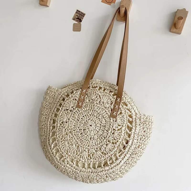 Sac En Paille Rond Ajouré Bandoulière – Solène Sac en paille rond ajouré avec bandoulière en cuir marron modèle Solène suspendu sur un mur blanc