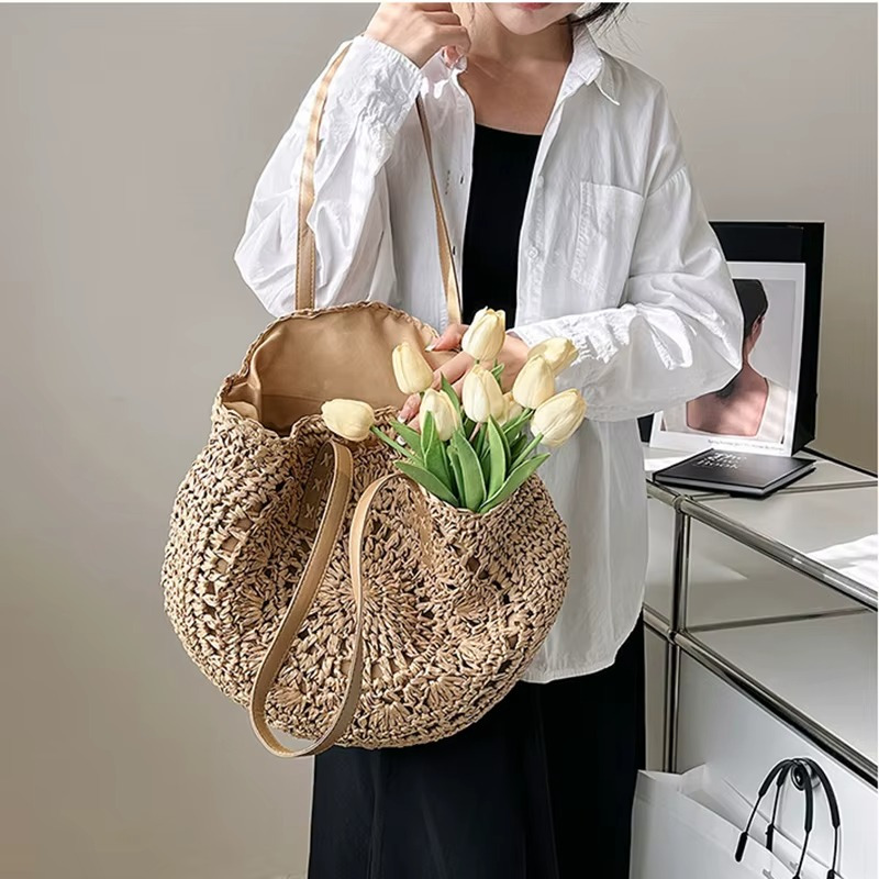 Sac En Paille Rond Ajouré Bandoulière – Solène Sac en paille rond ajouré avec bandoulière porté à l'épaule, modèle Solène, couleur naturelle, rempli de tulipes blanches