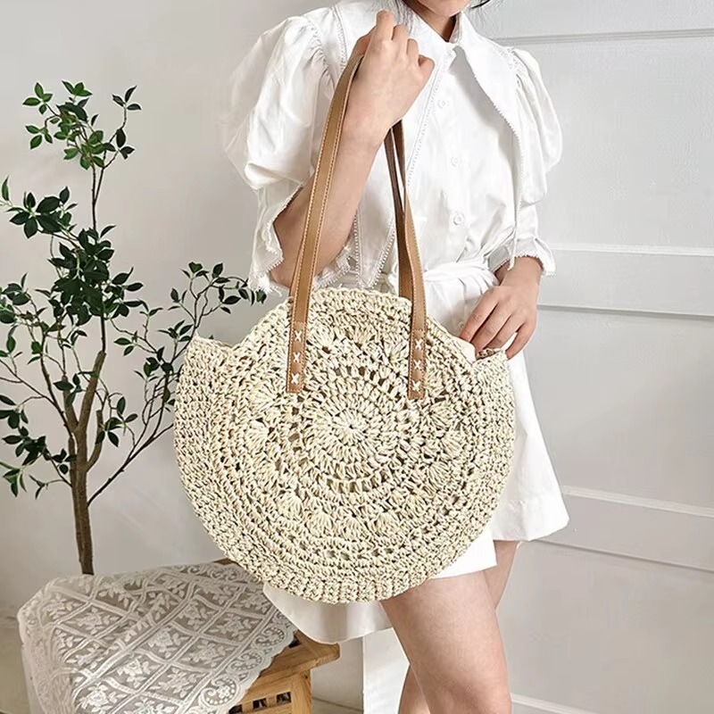 Sac En Paille Rond Ajouré Bandoulière – Solène Sac en paille rond ajouré beige avec bandoulière marron porté à l'épaule par une femme en robe blanche