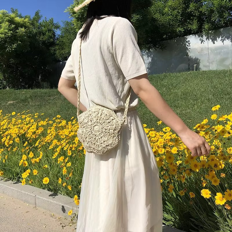 Sac en paille rond crochet dentelle plat couleur naturelle porté en bandoulière sur une femme habillée en blanc devant des fleurs jaunes