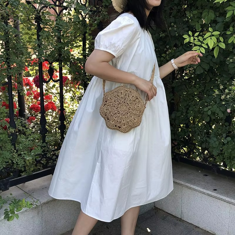 Sac en paille rond crochet dentelle plat couleur naturelle porté en bandoulière par une femme en robe blanche dans un jardin