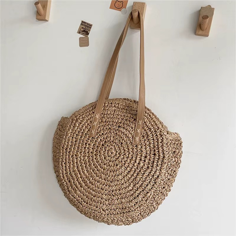 Sac En Paille Rond Tissage Uni Bandoulière – Adèle Sac en paille rond tissage uni avec bandoulière beige modèle Adèle suspendu sur un mur blanc