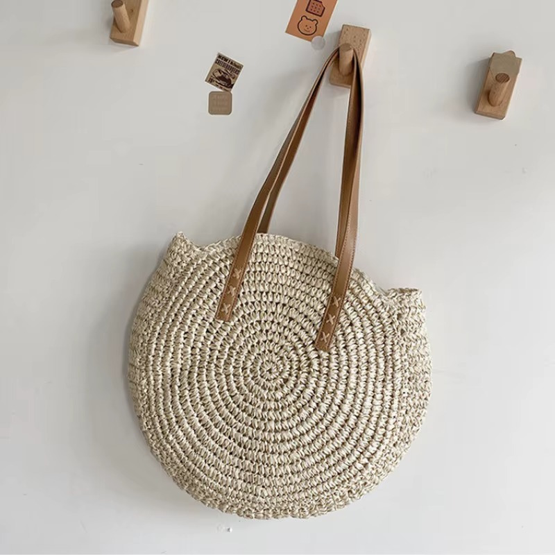 Sac En Paille Rond Tissage Uni Bandoulière – Adèle Sac en paille rond tissage uni beige avec bandoulière marron modèle Adèle suspendu au mur
