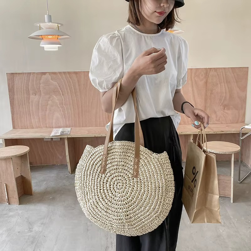 Sac En Paille Rond Tissage Uni Bandoulière – Adèle Sac en paille rond tissage uni avec bandoulière en cuir porté par une femme en intérieur