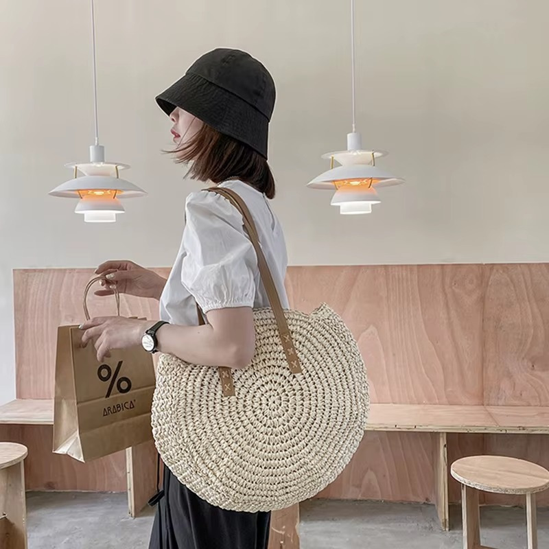 Sac En Paille Rond Tissage Uni Bandoulière – Adèle Sac en paille rond tissage uni avec bandoulière en cuir porté par une femme de profil dans un intérieur moderne