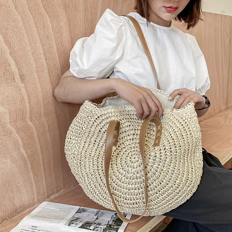 Sac En Paille Rond Tissage Uni Bandoulière – Adèle Sac en paille rond tissage uni couleur naturelle avec bandoulière en cuir porté par une femme en blouse blanche