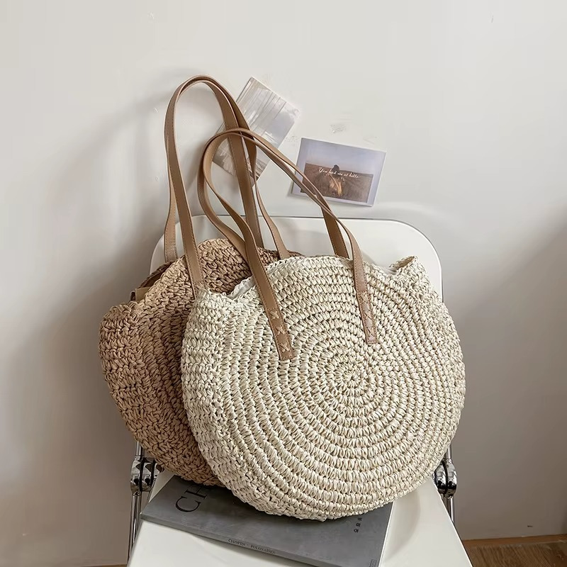 Sac En Paille Rond Tissage Uni Bandoulière – Adèle Sac en paille rond tissage uni beige avec bandoulière marron modèle Adèle posé sur une chaise blanche, vue de face