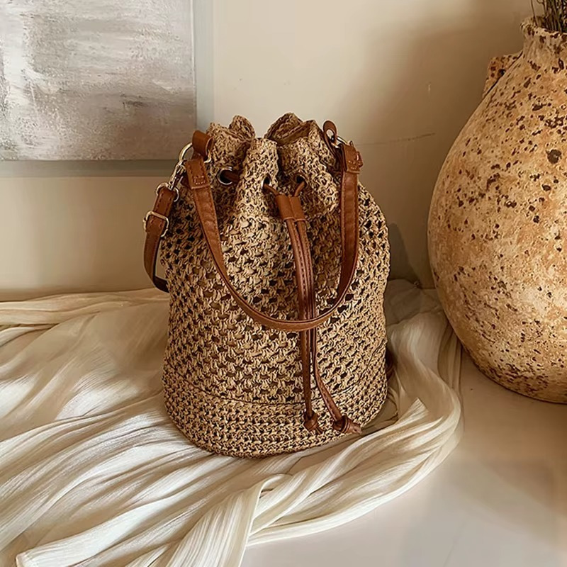 Sac en paille seau ajouré avec cordon coulissant et anses en cuir marron posé sur tissu blanc