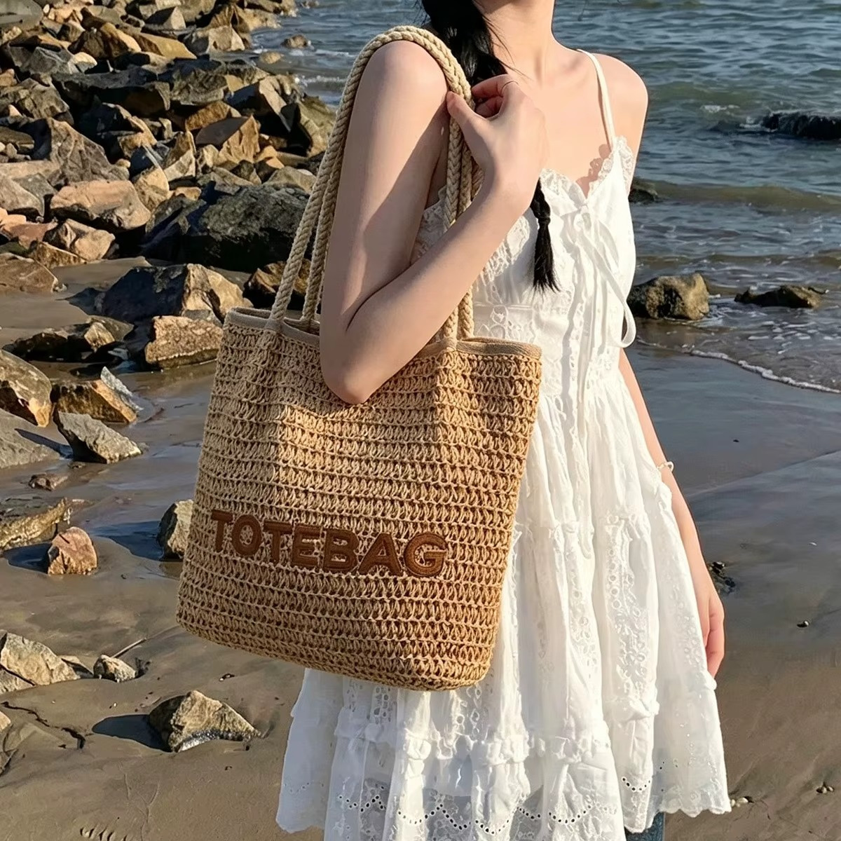 Sac En Paille Tressée Bandoulière – Paolita Sac en paille tressée bandoulière Paolita couleur naturelle avec inscription totebag porté sur l'épaule par une femme en robe blanche au bord de la mer