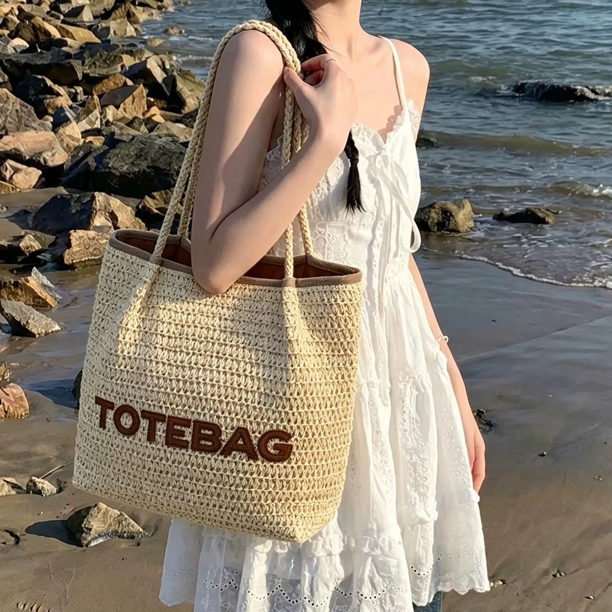 Sac En Paille Tressée Bandoulière – Paolita Sac en paille tressée bandoulière Paolita couleur naturelle avec inscription TOTE BAG marron porté par une femme en robe blanche sur la plage