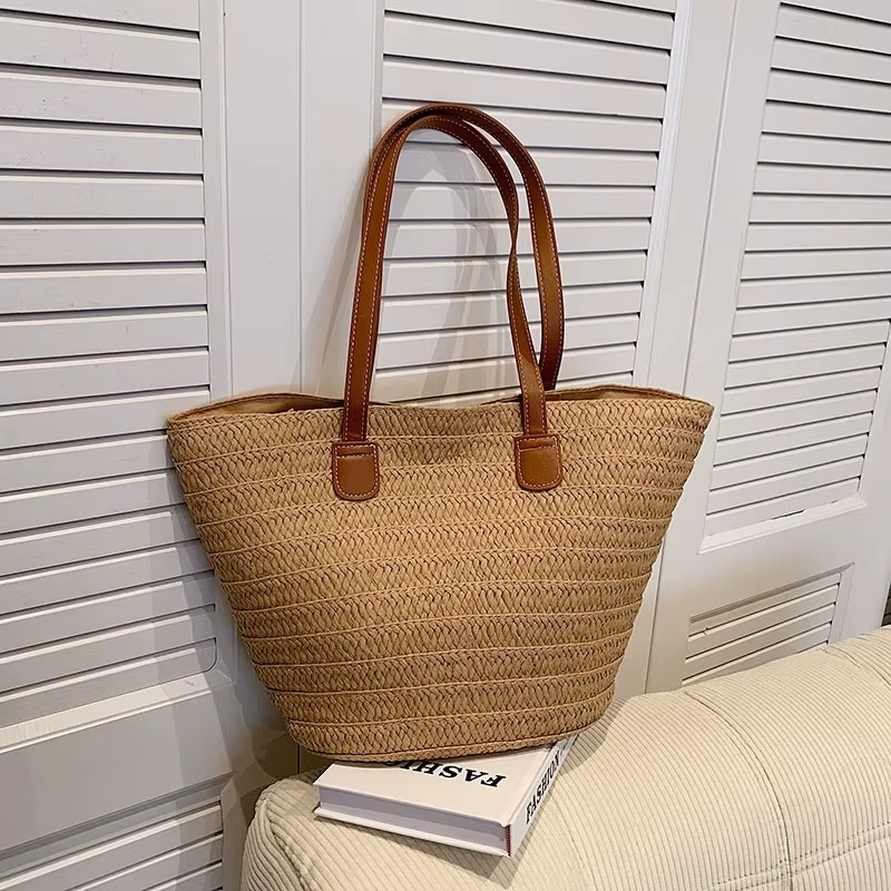 Sac en paille tressée bohème grande capacité modèle Océane avec anses en simili cuir marron posé sur un canapé clair devant un volet blanc