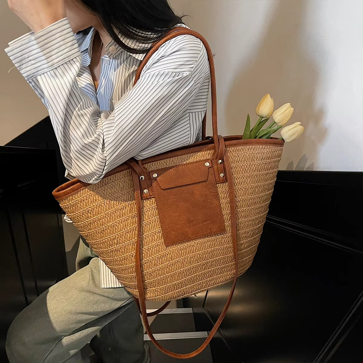 Sac en paille tressée cabas avec poche en cuir marron et rivets porté à l'épaule avec des tulipes à l'intérieur