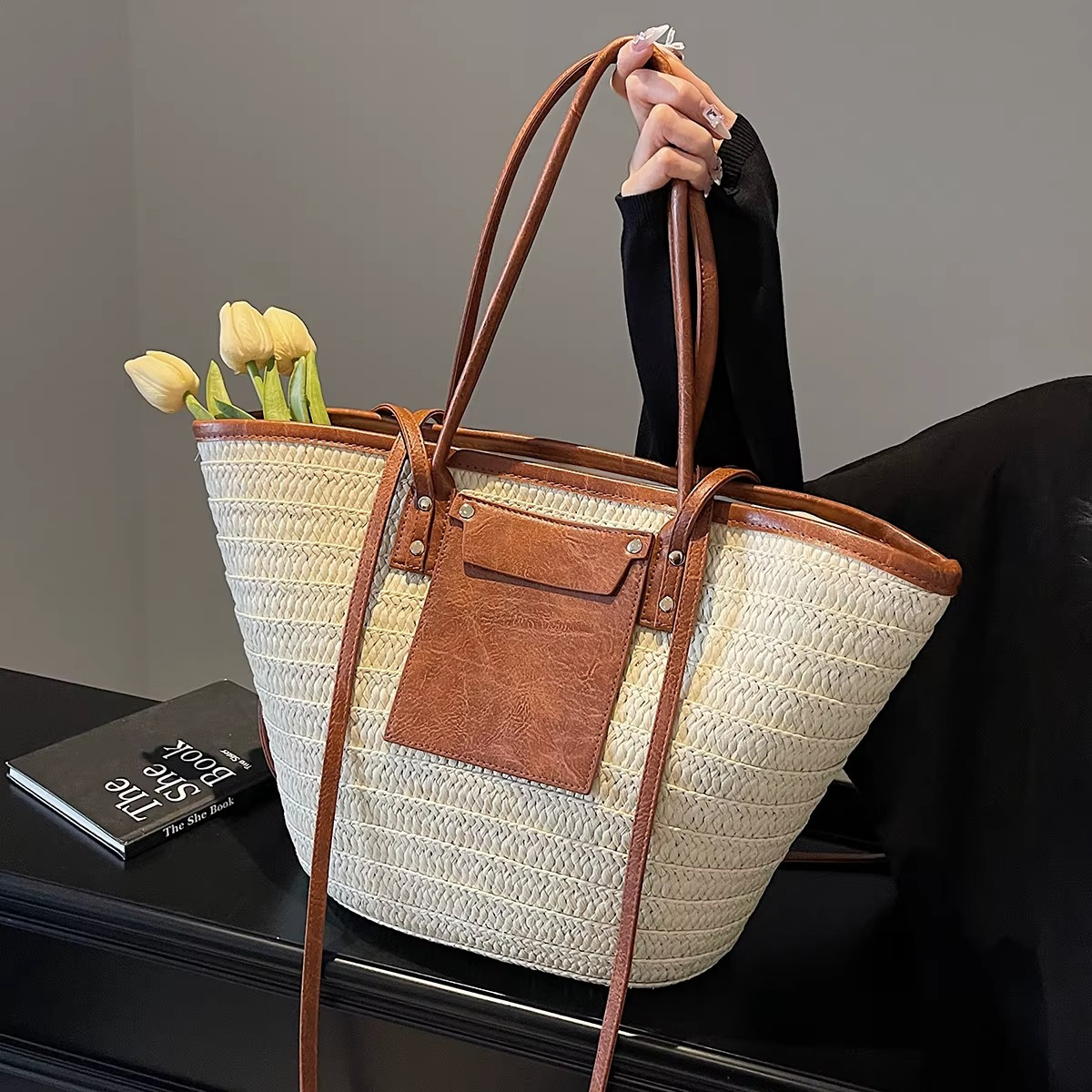 Sac en paille tressée cabas avec poche en cuir marron et rivets, modèle Constance, tenu par une main avec des tulipes jaunes à l'intérieur