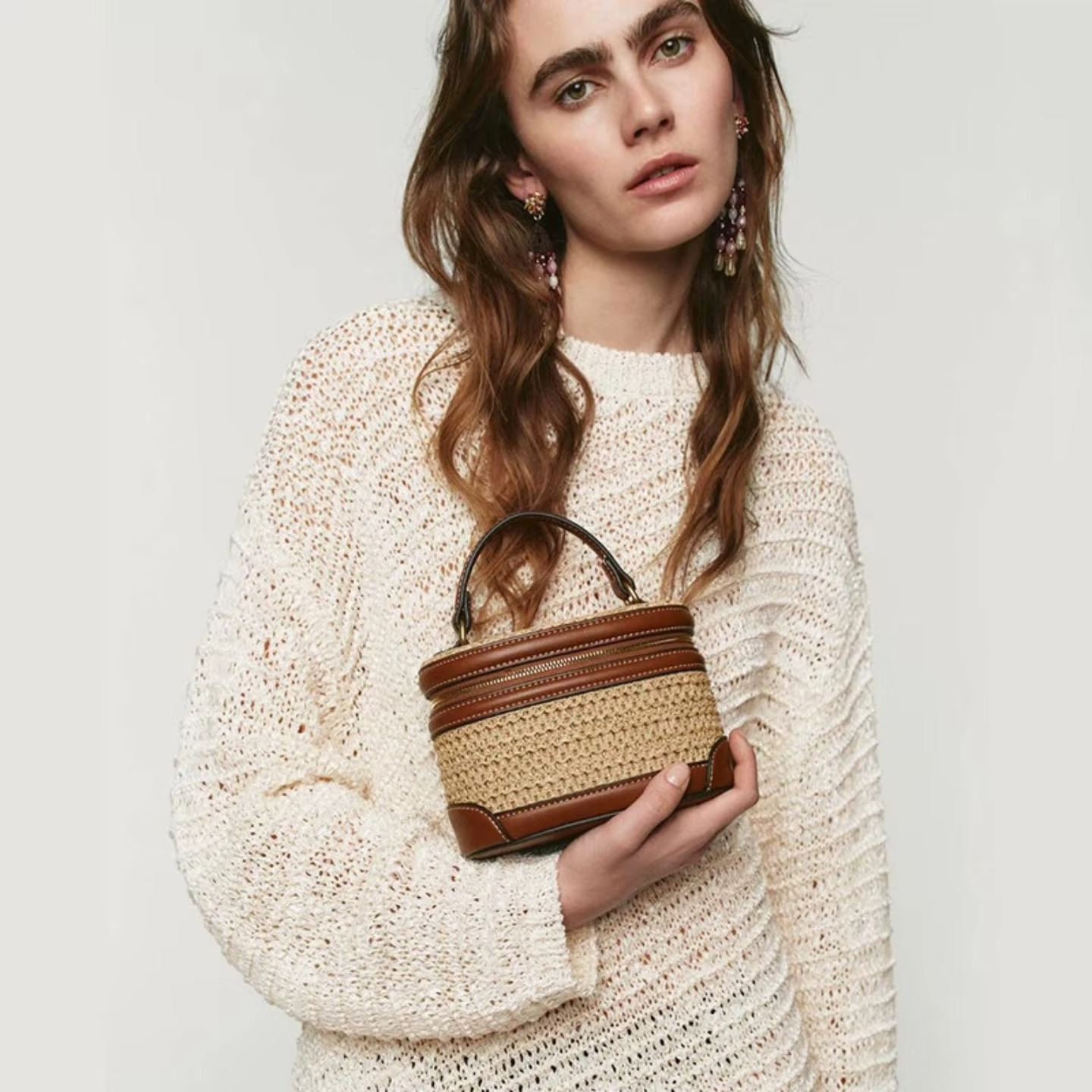 Sac en paille tressée mini boîte vintage Charlotte avec détails en cuir marron tenu par une femme en pull beige