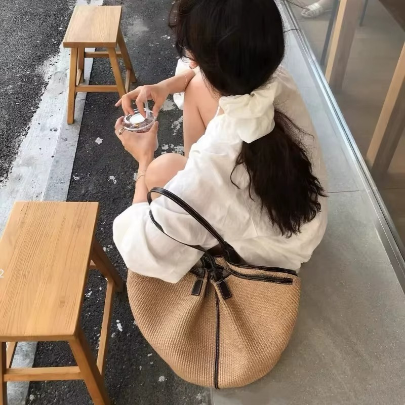 Sac en paille tressée seau avec liseré cuir noir porté sur l'épaule d'une femme assise de dos