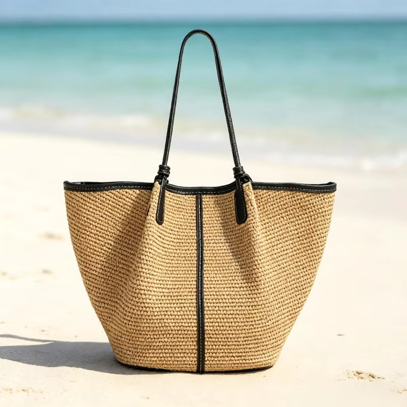 Sac en paille tressée seau avec liseré cuir noir, modèle Rosalinde, posé sur le sable face à la mer