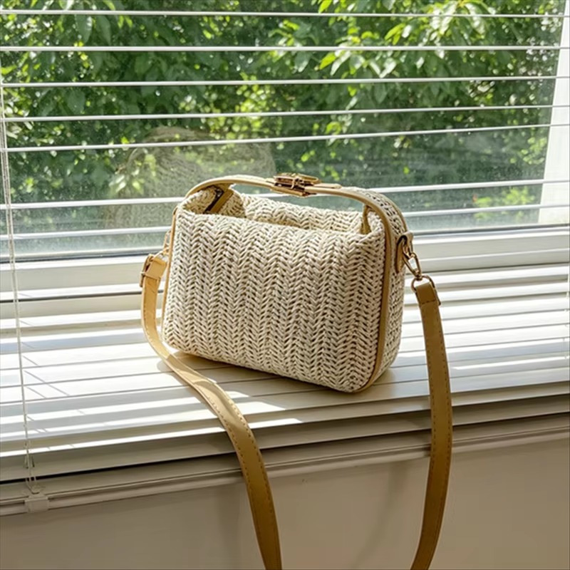 Sac en paille vintage rigide beige avec poignée et fermoir doré, bandoulière fine marron clair, posé sur un rebord de fenêtre devant des stores